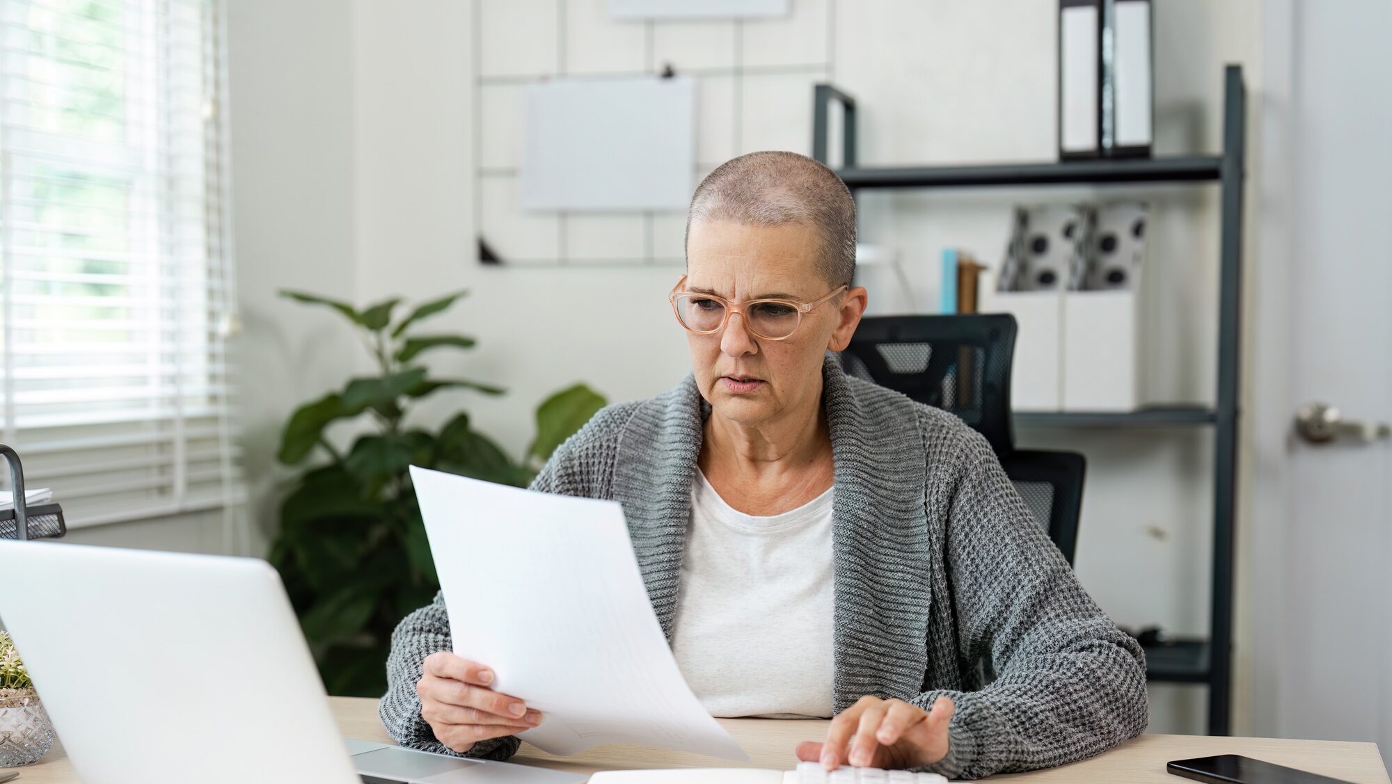 Senior Woman Reviewing Financial Documents at Home Office Desk with Laptop and Calculator